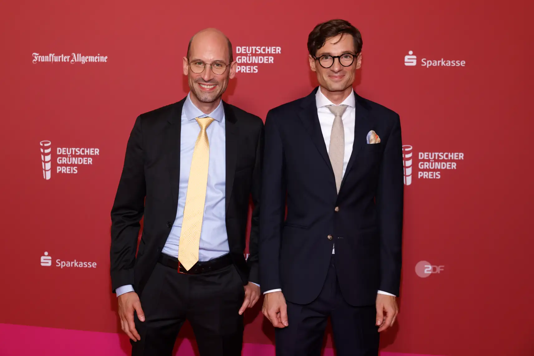 Two men in suits posing on a red backdrop with "Deutscher Gründer Preis" branding, a bald man on the left wearing a yellow tie and glasses, and a man on the right with dark hair and round glasses wearing a tan tie with a pocket square.