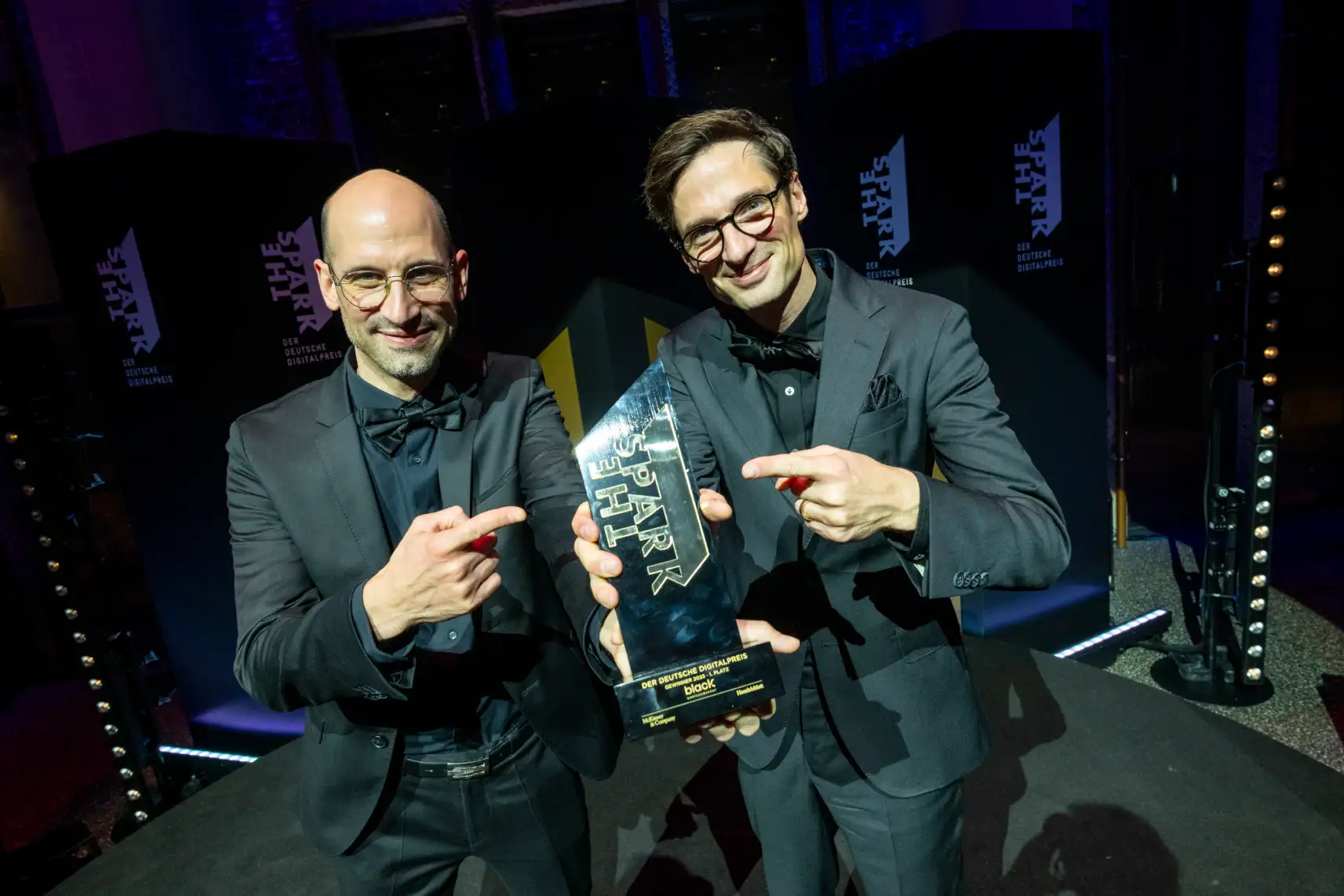 Two men in suits holding the "Der Deutsche Digitalpreis" award, pointing at it, smiling at the camera, standing on stage with "The Spark" logos and stage lighting in the background.